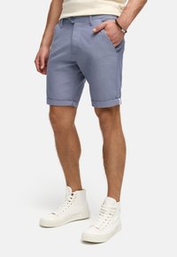 Himmelblaue Shorts mit umgeschlagenen Säumen, aus einem strukturierten Stoff. Vordertaschen und ein einzelner Knopfverschluss sind sichtbar. Getragen mit weißen High-Top-Sneakern.