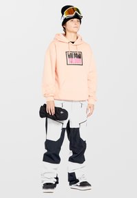 Pfirsichfarbener Hoodie mit einem schwarz-pinken Grafik-Logo, kombiniert mit schwarz-weißen Schneehosen, die Taschen und Reißverschlüsse haben, und hält schwarze Handschuhe.