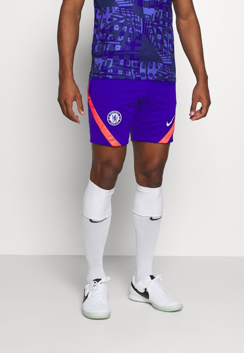 Homme portant un short de football Chelsea violet avec des bandes orange, des chaussettes blanches et des chaussures blanches Nike, debout sur fond blanc.