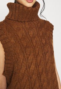 Hnědý rolákový svetr s kabelovým vzorem, bez rukávů a s výraznou texturou, zakončený žebrovaným lemem. Pohodlný a teplý vzhled.