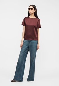 Μπλούζα μαρόν σατέν με κοντά μανίκια σε συνδυασμό με μπλε τζιν παντελόνι φαρδύ με ευρεία πόδια· μοντέλο που φορά σκοτεινά γυαλιά ηλίου, στέκεται μπροστά σε λευκό φόντο.