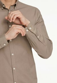 Cămașă bej deschis, cu nasturi, cu guler. Are nasturi albi și mâneci lungi cu manșete cu un singur nasture. Textură netedă.