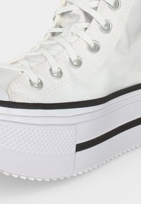 Gros plan sur une basket montante blanche avec œillets argentés, lacets blancs et semelle plateforme blanche épaisse avec détail de rayure noire.