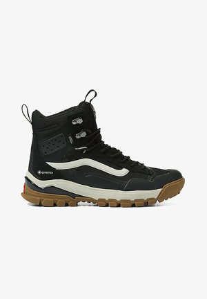 Czarny wysokogórski but turystyczny z brązową, ryflowaną podeszwą, białym bocznym paskiem, metką Gore-Tex, metalowymi haczykami na sznurówki i wyściełanym wsparciem kostki.