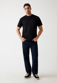 Zwarte t-shirt met korte mouwen met een subtiele, reliëflogo patroon op de voorkant, gecombineerd met donkerblauwe jeans en zwarte sneakers.