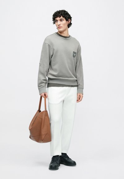 Sweatshirt cinzenta com um remendo, calças brancas feitas à medida e uma bolsa castanha em couro com textura. Ténis pretos completam o conjunto.