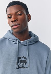 Jasnoniebieska bluza z kapturem ze sznurkami, ozdobiona czarnym logo graficznym i napisem na piersi. Miękka tkanina i luźny krój.