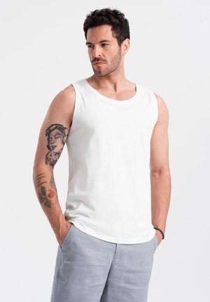 Débardeur en coton blanc avec un col rond et de larges ouvertures pour les bras, porté par un homme, assorti à un short gris clair. Présente des tatouages visibles sur le bras.