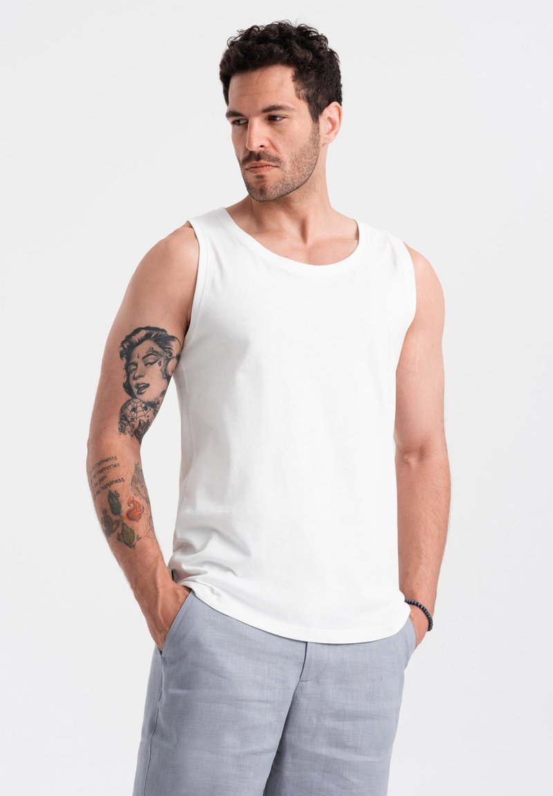 Débardeur en coton blanc avec un col rond et de larges ouvertures pour les bras, porté par un homme, assorti à un short gris clair. Présente des tatouages visibles sur le bras.