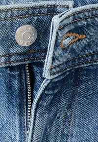 Gros plan d'un jean en denim bleu non fermé avec un bouton argenté étiqueté "MNG JEANS" et des coutures brunes visibles.