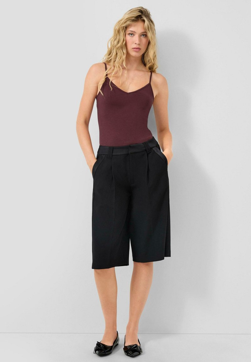 Kobieta z długimi falistymi blond włosami, ubrana w bezrękawnik w kolorze bordowym, czarne szorty do kolan o szerokim kroju oraz czarne płaskie buty, stojąca przy jasnej ścianie.