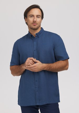 Mężczyzna o krótkich brązowych włosach i brodzie, ubrany w niebieską koszulę z krótkim rękawem i guzikami, stojący na jednolitym jasnym tle.