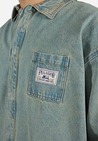 Camisa de mezclilla azul claro con cuello puntiagudo, bolsillo delantero con una etiqueta 'PEGADOR', detalles de costura visibles y textura ligeramente desgastada.