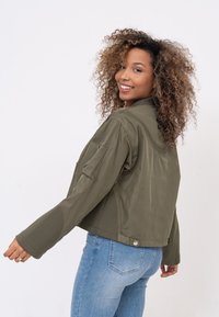 Olivgrüne, kurz geschnittene Jacke aus leichtem Stoff mit Seitentaschen und Reißverschlussdetails, kombiniert mit blauen Jeans. Strukturierte Ärmelbündchen.