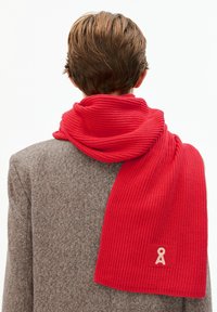 Persoon gezien van achteren, gekleed in een tan kleurige jas en een felrode gebreide sjaal die over de schouders hangt met een klein beige logo.