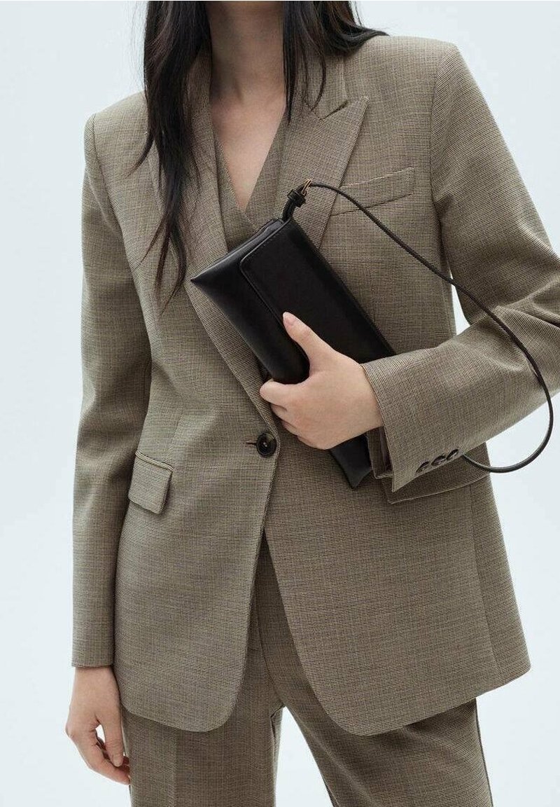 Personne portant un costume beige tenant une pochette noire rectangulaire avec une lanière de poignet sur un fond clair uni.
