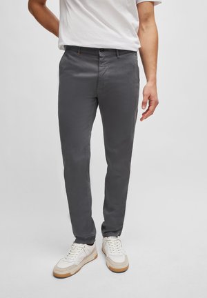 Adulte portant un pantalon slim gris foncé, un t-shirt blanc et des baskets blanches à semelles beiges, debout devant un fond uni.