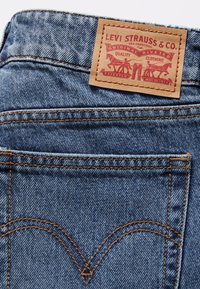 Jeans en denim bleu avec une patch en cuir tan portant l'inscription "LEVI STRAUSS & CO." au dos, avec des coutures orange et un design classique de poche arrière.