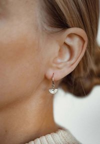 Boucle d'oreille en métal avec un pendentif en forme de éventail, arborant un motif texturé et un petit accent rond en haut. La couleur semble être argent.