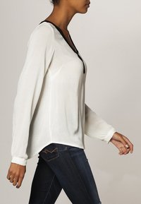 Blouse blanche à manches longues avec un tissu texturé, dotée d'un col orné de noir et d'une coupe décontractée, portée avec un jean denim foncé.