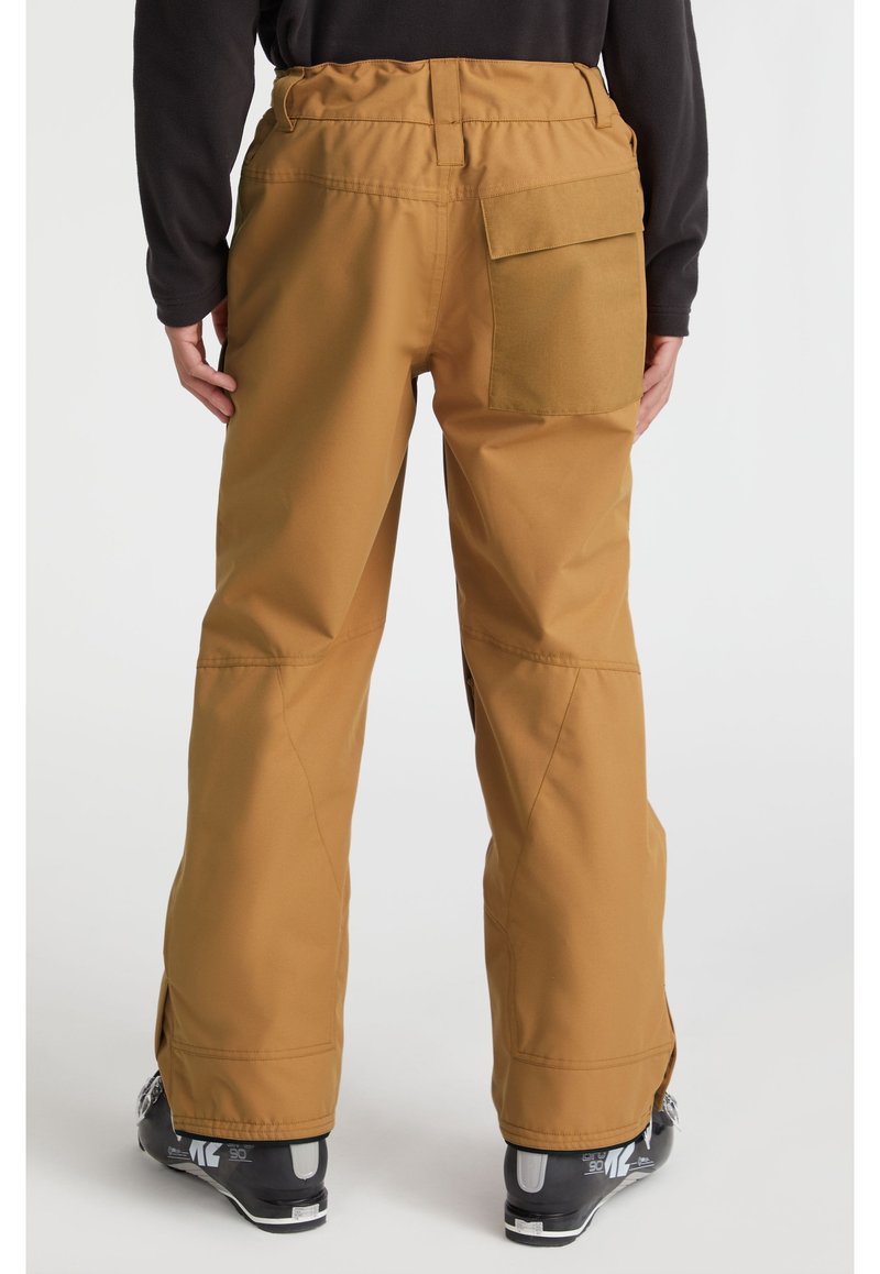 Person in beigen Schneehosen mit einer großen hinteren Tasche, schwarzem Oberteil und schwarzen Skischuhen vor einem weißen Hintergrund.