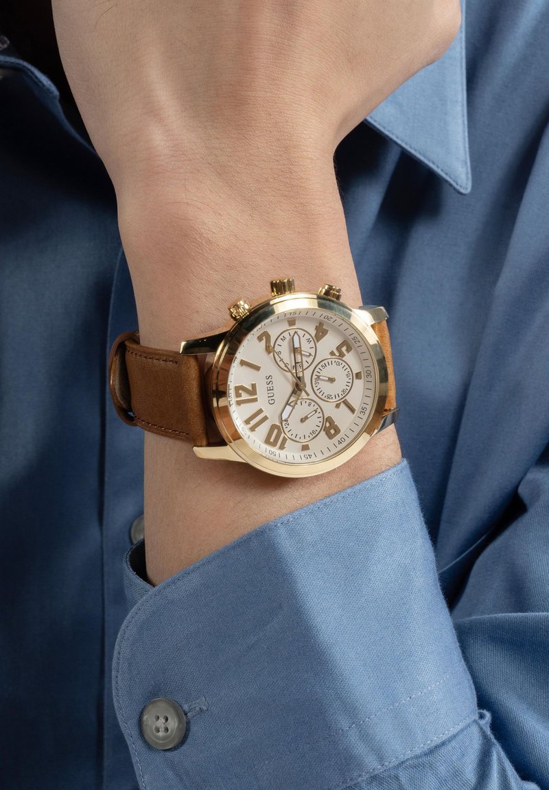 Orologio con cinturino in pelle marrone, quadrante bianco, cassa dorata, grandi numeri e tre sotto-quadranti. Camicia azzurra con colletto sullo sfondo.
