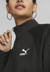 Schwarze Zip-Jacke mit geripptem Kragen, ausgestattet mit dem weißen Puma-Logo. Glattes Material, taillierte Passform, mit kontrastierenden Schulterakzenten.