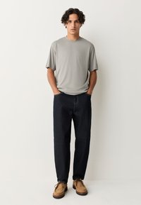 Gray short-sleeve t-shirt, dark blue straight-leg jeans, and light brown shoes. Model stands with hands in pockets against a plain background.