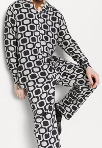 En person klädd i en matchande svart och vit geometriskt mönstrad skjorta med knappar och byxor, sitter med ett knä upphöjt, mot en vit bakgrund.