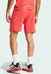 Shorts de sport rouges, en tissu léger, coupe ample. Chaussettes de sport blanches et baskets noires/rouges avec des rayures distinctes. Texture lisse.