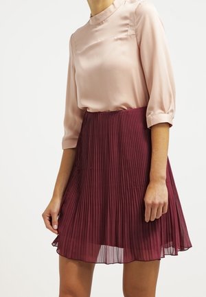 Lichtroze satijnen blouse met driekwartmouwen, gecombineerd met een bordeauxrode geplooide rok die boven de knie uitkomt en een gevarieerd patroon toont.