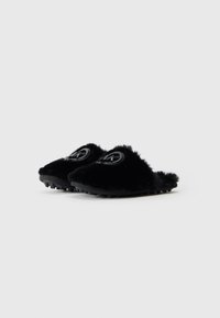 Schwarze, flauschige Slipper mit Gummisohlen und einem runden, glänzenden Logoemblem oben, gezeigt vor einem schlichten weißen Hintergrund.