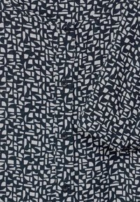 Camicetta bianca e nera con motivo geometrico, con bottoni davanti, maniche corte e una texture di tessuto leggero.