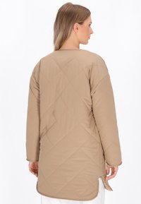 Cappotto lungo beige trapuntato con scollatura rotonda, fessure laterali e tessuto morbido, che presenta un design semplice e minimalista.