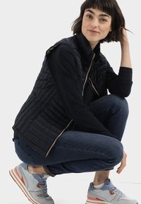 Marineblå quiltet vest med høj krave, parret med en sort langærmet skjorte og blå jeans. Funktioner sporty grå sneakers med orange detaljer.