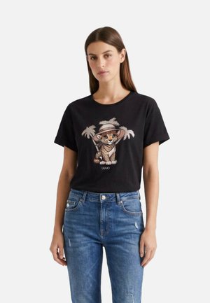 Donna che indossa una maglietta nera con un disegno di un ghepardo cartoon con un cappello e palme, abbinata a jeans blu su uno sfondo bianco.