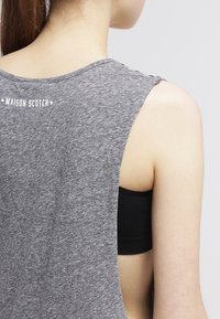 Grå ärmlös topp med öppen ryggdesign, tillverkad av mjukt, texturerat tyg och med en vit logotyp på övre delen av ryggen. Svart bh synlig under. 
