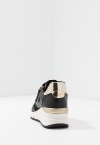 Černé tenisky s texturovanou kůží, kovovými zlatými akcenty a bílou podrážkou. Mají zipový uzávěr a kulaté logo na zadní straně.