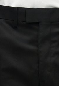 Κοντινό πλάνο της μέσης μαύρου παντελονιού με θηλιές για ζώνη και κρυφό φερμουάρ, με λευκό φόντο.