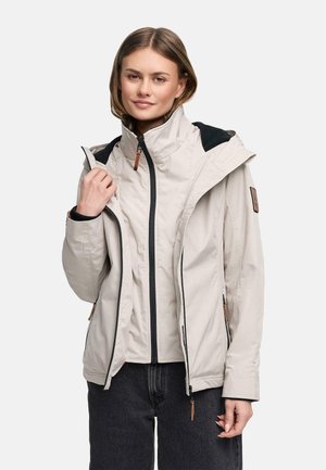 Jeune femme portant une veste à capuche beige clair par-dessus une couche intérieure zippée, associée à un pantalon foncé, debout devant un fond uni.