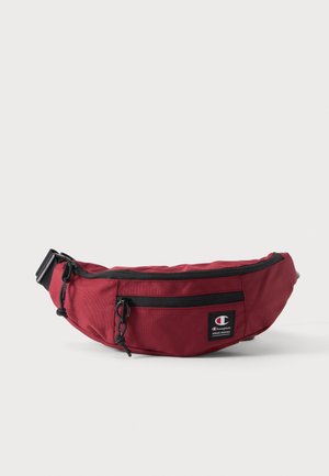 Rode Champion-heuptas met zwarte ritsen, een verstelbare riem en een logopatch aan de voorkant, getoond tegen een egale witte achtergrond.