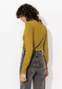 Pull à manches longues couleur moutarde avec une coupe ajustée, associé à un jean gris taille haute comportant deux poches arrière et de subtils accents de boutons argentés.