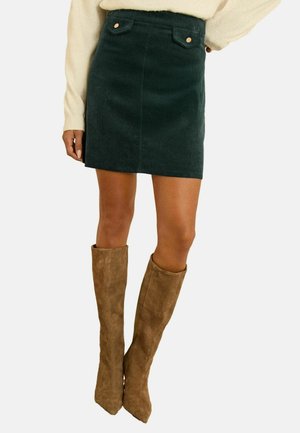 Grüne Cordmini mit Fronttaschen und Metallknöpfen, kombiniert mit beigen kniehohen Wildlederstiefeln. Glatte Textur und taillierte Passform.