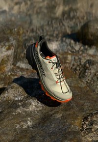 Ελαφρύ γκρι αθλητικό παπούτσι με μαύρες λεπτομέρειες και πορτοκαλί σόλα. Διαθέτει άνω μέρος από πλέγμα και σύστημα δέσιμο για σταθερότητα.