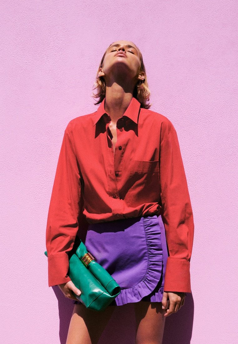 Mujer con camisa roja y falda morada que sostiene un bolso de mano verde, de pie contra una pared texturizada de color rosa brillante con la cabeza inclinada hacia atrás.