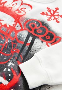 Vit sweatshirt med en grafisk design i röd och svart, med mönster som inkluderar snöflingor och abstrakta former på en texturerad bakgrund.