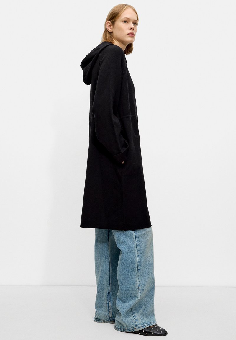 Manteau à capuche noir au fit décontracté, à manches longues et avec des poches latérales, associé à un jean en denim bleu ample et des chaussures noires à clous.