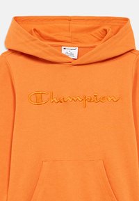 Πορτοκαλί φούτερ με κουκούλα, κατασκευασμένο από απαλό ύφασμα. Διαθέτει κεντητό λογότυπο "Champion" στο ίδιο πορτοκαλί χρώμα. Περιλαμβάνει μπροστινή τσέπη.