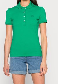 Chemise polo verte en coton avec un col classique, des manches courtes et une patte de boutonnage à trois boutons. Présente un petit logo brodé sur la poitrine.