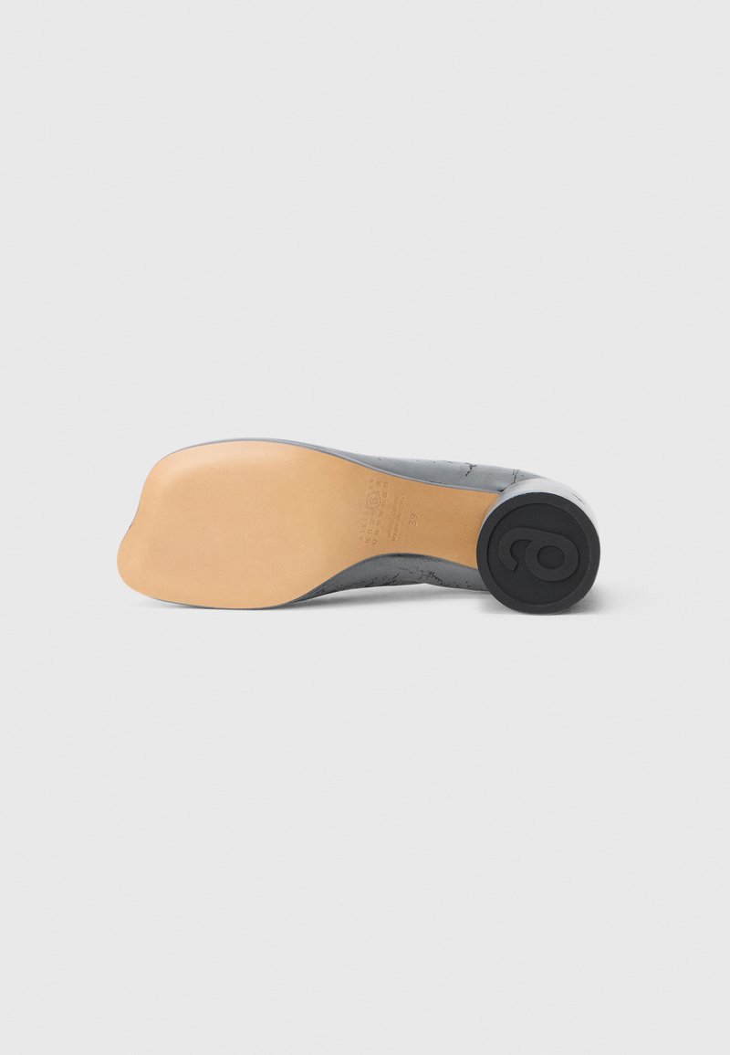Chaussure grise pour femme couchée sur le côté montrant une semelle beige lisse et un talon noir rond avec un design en relief sur fond uni.